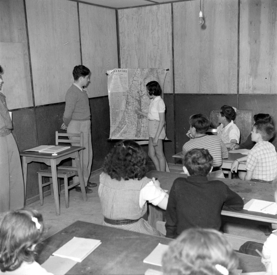 Leerlingen en een leerkracht voor een landkaart in een leslokaal van het kinderd, Bestanddeelnr 255-0519