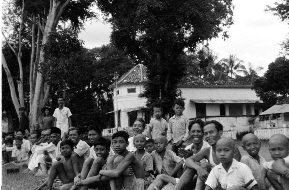 Lachende Indonesische jongens en mannen zitten op een terrein voor een koloniaal, Bestanddeelnr 5441