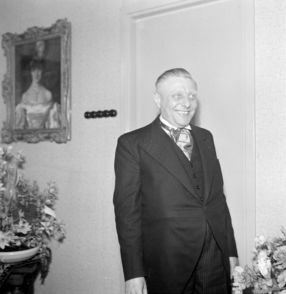 Man in gelegenheidskleding, op de achtergrond een portret van een vrouw, Bestanddeelnr 255-8520