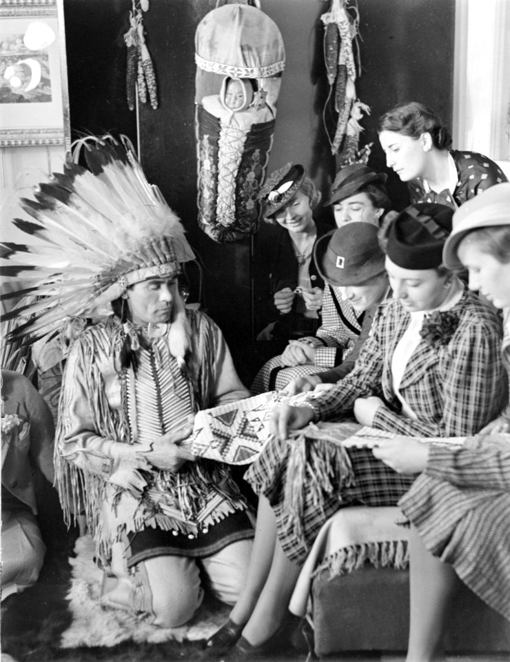 Man in Indianenpak bekijkt Indiaans handwerk met zes vrouwen, de meesten met een, Bestanddeelnr 190-0776