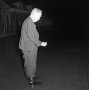 Man in kostuum staat met laarzen in laag water in een straat en houdt een vierka, Bestanddeelnr 900-8367