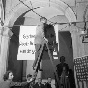 Man hangt spandoek op met de tekst 'Geschenn de Roode Kruis organisati, Bestanddeelnr 900-8202