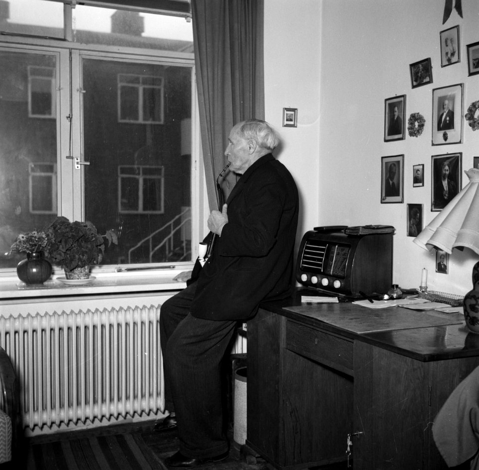 Man met een pijp staat tegen een bureau geleund voor het raam te roken, Bestanddeelnr 252-9126