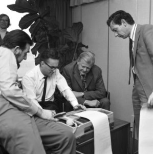 Mannelijke kantooremployees staan samen gebogen over een teletypewriter fax, Bestanddeelnr 167-1016