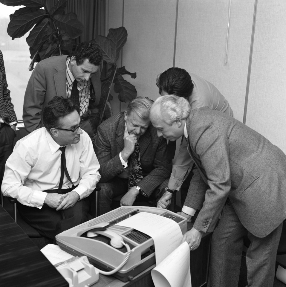 Mannelijke kantooremployees staan samen gebogen over een teletypewriter fax, Bestanddeelnr 167-1019