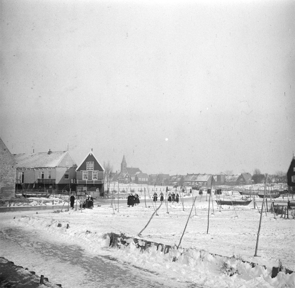 Mensen in klederdracht lopen door de sneeuw langs een rij huizen, Bestanddeelnr 254-1939