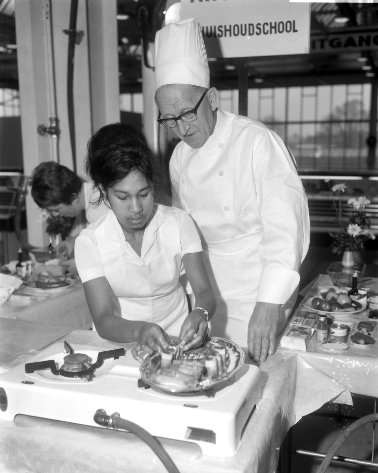 Meisje bezig met het bereiden van schotels onder toeziend oog van chef-kok H. E…, Bestanddeelnr 918-4641