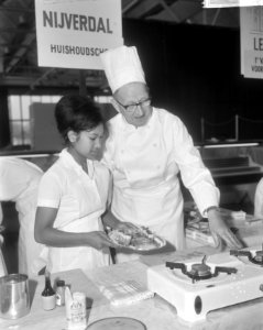 Meisje bezig met het bereiden van schotels onder toeziend oog van chef-kok H. E…, Bestanddeelnr 918-4640
