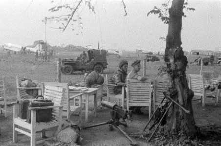 Militairen zitten op stoelen op een vliegveld. Om hen heen wapens en aktetassen, Bestanddeelnr 2231