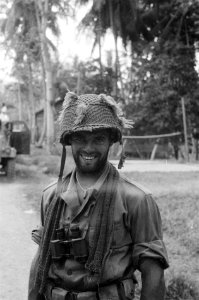 Militair met helm poseert. Achter hem een volleybalveld, Bestanddeelnr 3249