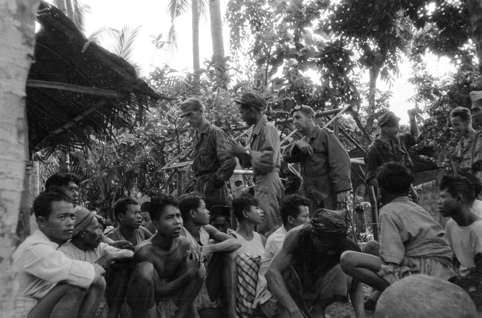 Operatie Schoonschip. Groep Indonesische mannen zitten op de grond voor een hut., Bestanddeelnr 5398