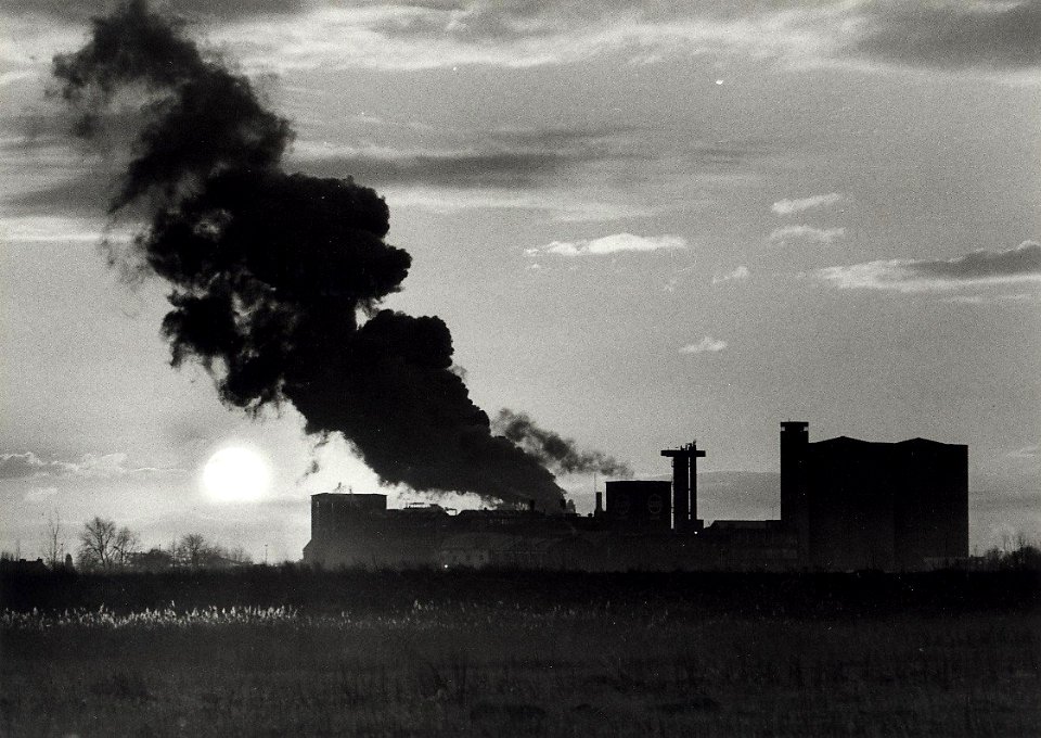 Tegen het licht van de lage zon lijkt de suikerfabriek in Halfweg inkt zwarte wolken uit te blazen. Aangekocht in 1977 van fotograaf C. de Boer. Identificatienummer 54-005105, NL-HlmNHA 1478 25900 K 38