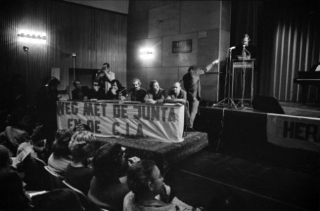 Toeschouwers bij podium, waarop door middel van een spandoek wordt geprotesteerd, Bestanddeelnr 926-9629