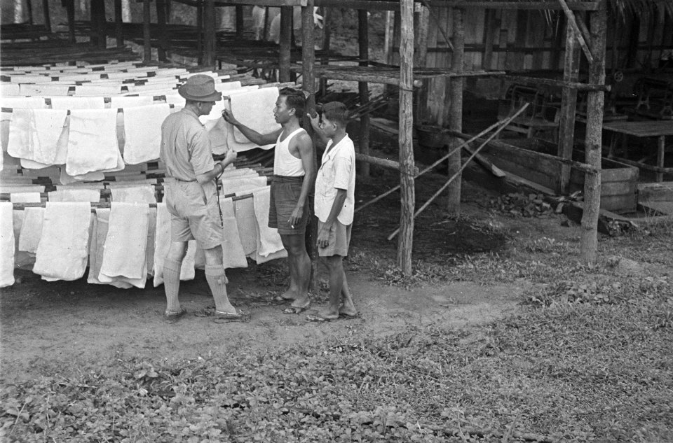 Twee jonge Indonesische mannen tonen een man in uniform met hoed een wasserij, Bestanddeelnr 122-5-2