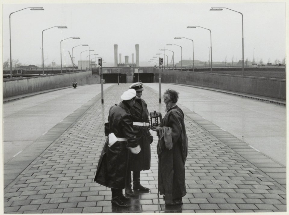 Twee agenten bij de ingang van de Velsertunnel in gesprek met een verklede man met een zandloper symbool van kortstondigheid. NL-HlmNHA 54010737