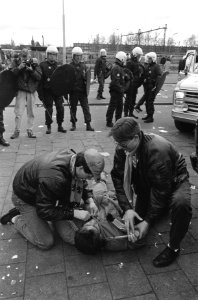 Twee mannen verzorgen een gewonde, op de achtergrond politie, Bestanddeelnr 934-4449
