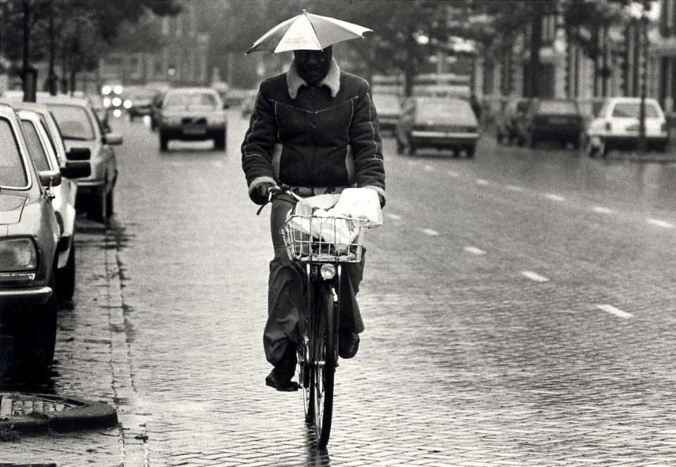 Onder moeders paraplui', Norris Bennet (folk-muziekant) op de fiets. Aangekocht in 1987 van United Fotos de Boer bv. - Negatiefnummer 25593 k 24. - Gepubliceerd in het Haarlems Dagblad van 09.10.1985
