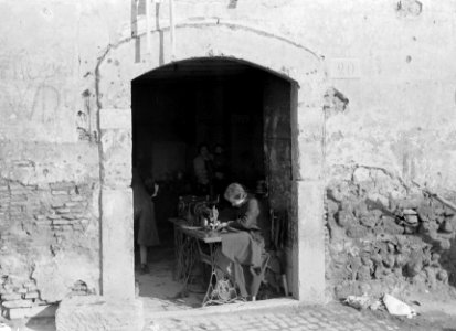 Vrouw zit in een deuropening achter een naaimachine met op de achtergrond een vr, Bestanddeelnr 191-1258