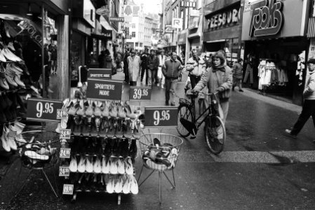 Vrouw met fiets aan de hand bekijkt voor een winkel uitgestalde schoenen, Bestanddeelnr 932-5159