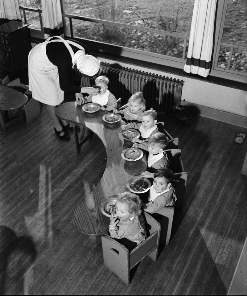Zes peuters zitten aan een halfronde tafel te eten. Een wordt gevoerd door een v, Bestanddeelnr 901-4153
