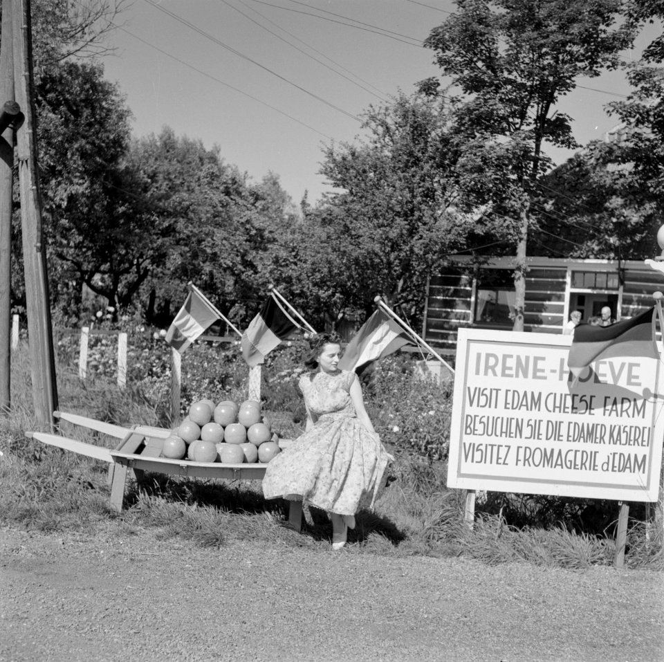 Volendam Irene-Hoeve meisje zit op een berrie met kazen, naast een reclamebord, Bestanddeelnr 254-3998