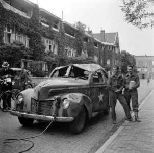 Vernielde legerauto wordt gesleept, rechts poseren twee militairen, Bestanddeelnr 900-3202