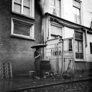 Vrouw met paraplu kookt wasgoed op een buitenfornuis. Het gebouw rechts is een , Bestanddeelnr 900-6398