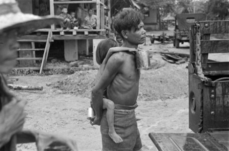 Indonesische man met kind op de rug. Kind houdt een blik vast. Jeeps zijn te zie, Bestanddeelnr 124-2-3