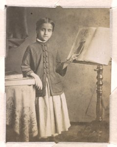 Adelina Patti, full-length studio portrait, facing front, standing, with a table on the left and a music stand on the right, pointing with her left hand at a portrait of Jenny Lind printed LCCN2015651565