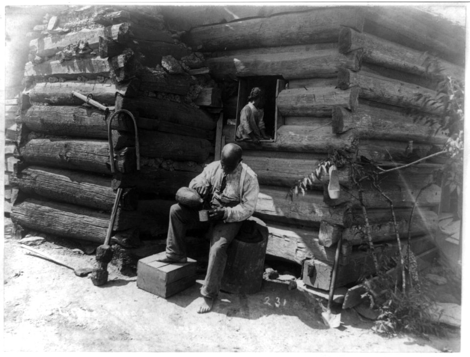 Afro-American man seated outside log cabin, pouring drink into cup and girl seated in window of the cabin LCCN92506349