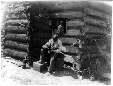 Afro-American man seated outside log cabin, pouring drink into cup and girl seated in window of the cabin LCCN92506349