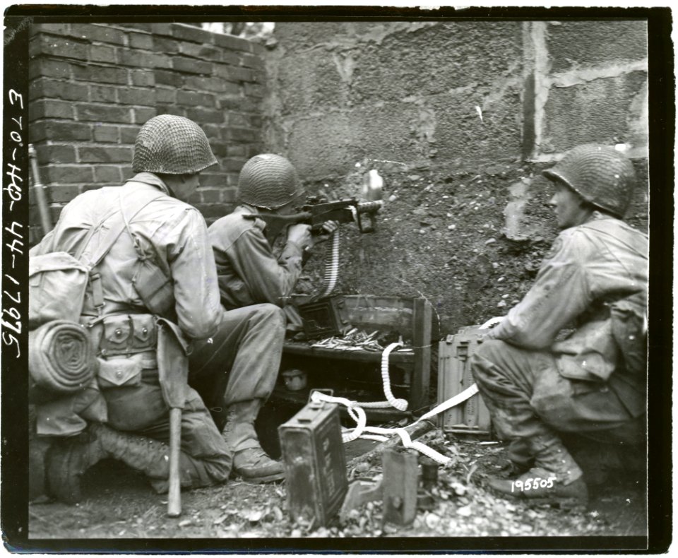 111-SC-195505 - PFC Victor Henry, Pontotoc, Miss., fires his machine gun through a hole in a wall, at Germans in a barn 300 yards away, beyond Kohlscheid, Germany. He is flanked by two of his buddies