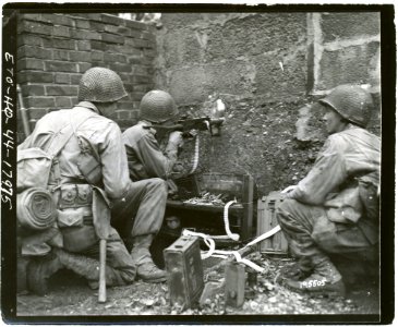 111-SC-195505 - PFC Victor Henry, Pontotoc, Miss., fires his machine gun through a hole in a wall, at Germans in a barn 300 yards away, beyond Kohlscheid, Germany. He is flanked by two of his buddies
