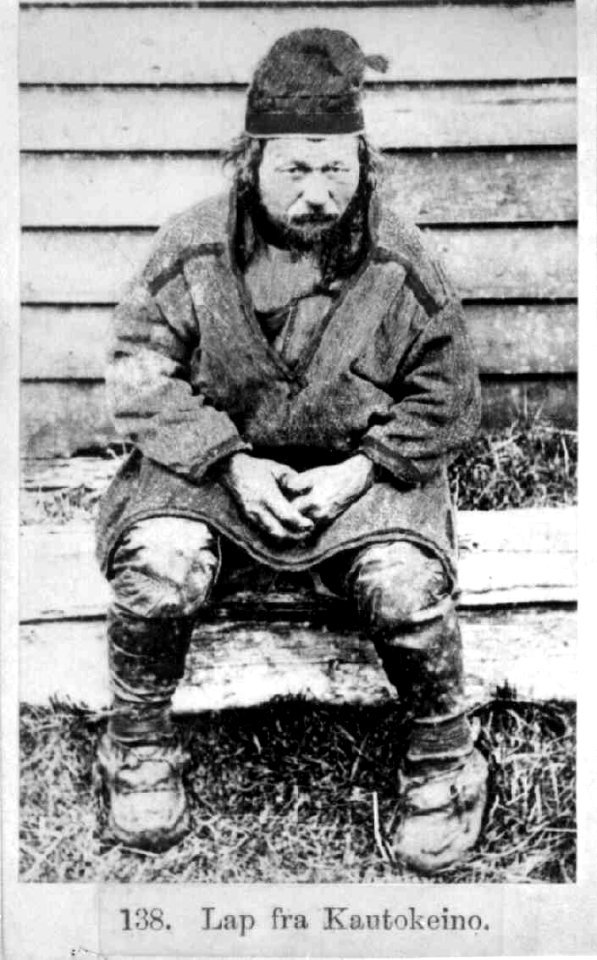 138. Mannsdrakt, Kautokeino (s-h). Mann sittende utendørs foran trevegg - Norsk folkemuseum - NF.06145-004