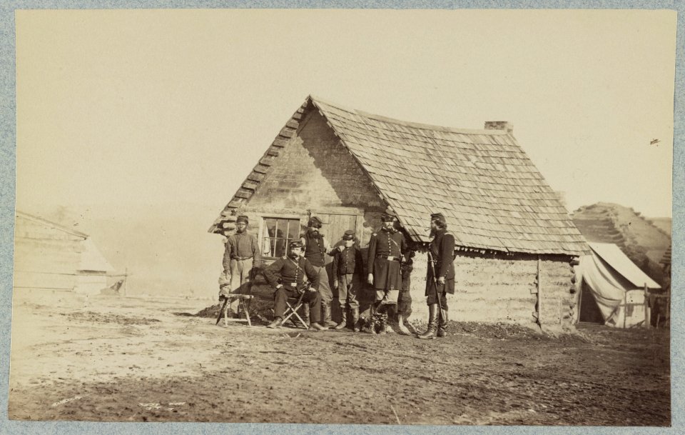 A group of soldiers, and two young men, one an African American, stand outside of log cabin quarters LCCN2013647883