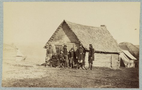 A group of soldiers, and two young men, one an African American, stand outside of log cabin quarters LCCN2013647883