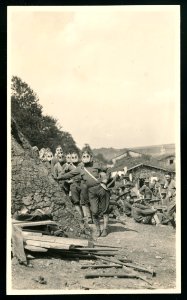 A line of soldiers posed, facing front, standing next to earthenwork fortifications, wearing gas masks over their faces, other soldiers sit on the ground nearby LCCN2016645678