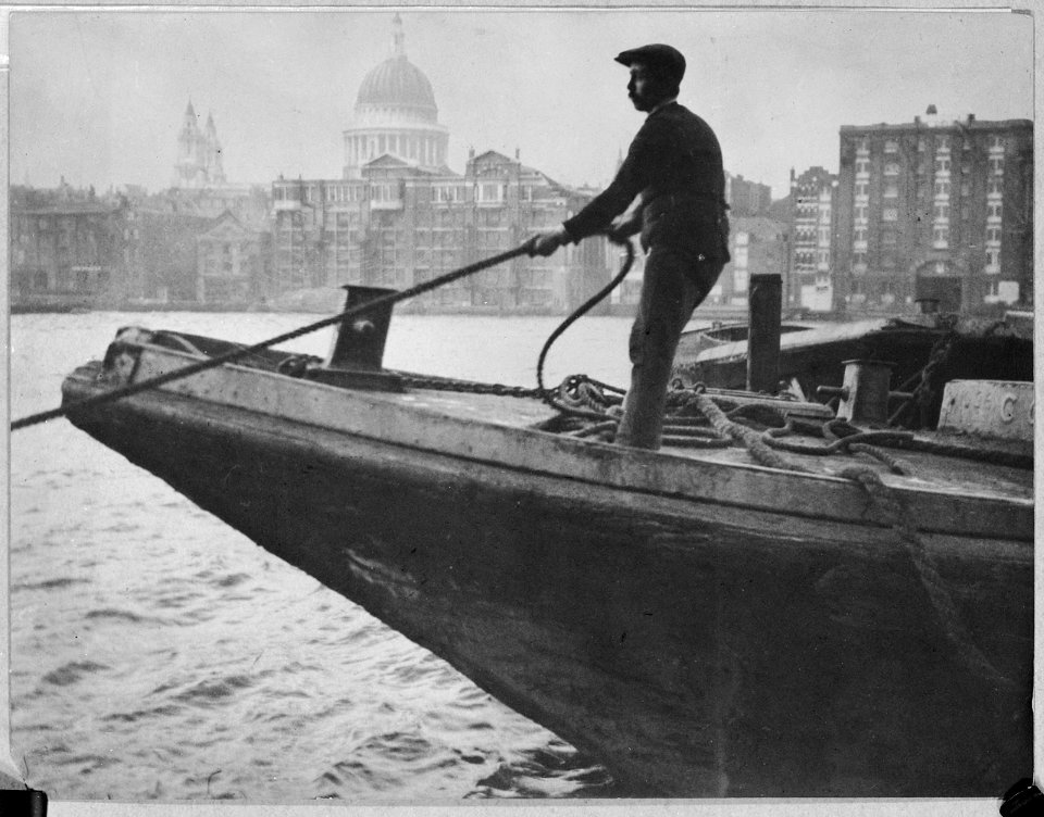 A lighterman standing on the bow of a lighter, hauling on a rope. RMG L8581
