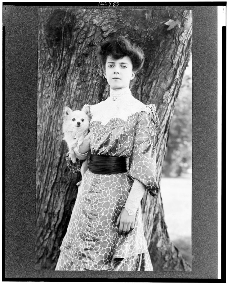 Alice Roosevelt Longworth, three-quarter length portrait, standing in front of tree, facing slightly left, holding small dog LCCN99403687