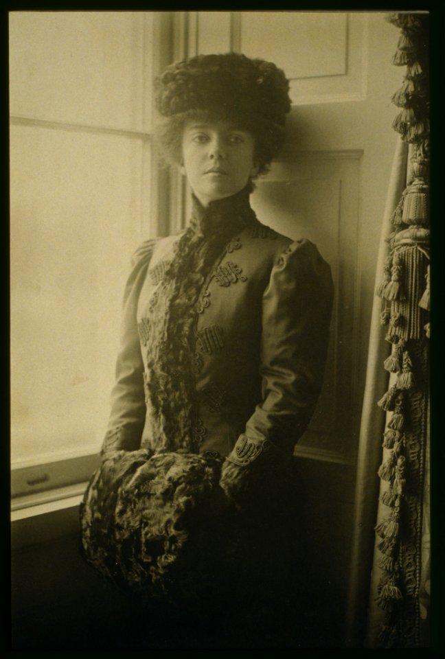 Alice Roosevelt Longworth, three-quarter length portrait, wearing hat and muff, standing at window, facing front LCCN2001697085