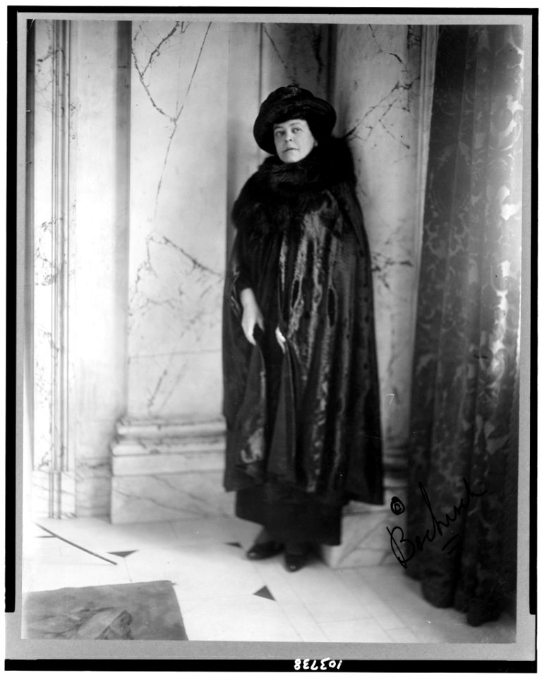 Alva E. Belmont, full-length portrait, standing, leaning against column, facing slightly left dressed in cape with fur neckpiece, hat LCCN91787573