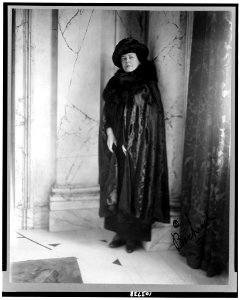 Alva E. Belmont, full-length portrait, standing, leaning against column, facing slightly left dressed in cape with fur neckpiece, hat LCCN91787573