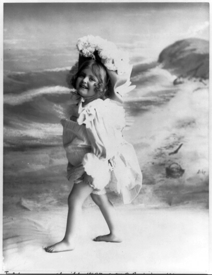 Barefoot young girl wearing dress and hat; fake beach scenery in background LCCN2002711742