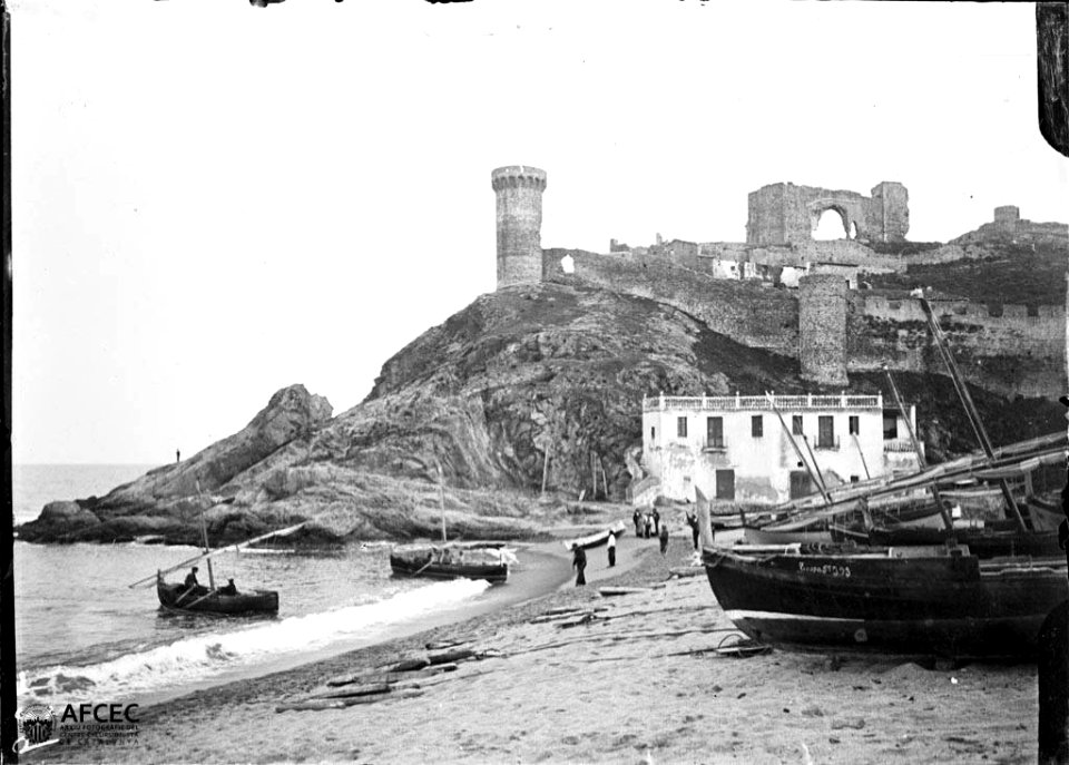 Barques i gent a la platja de Tossa de Mar amb un edifici i part de la muralla al fons