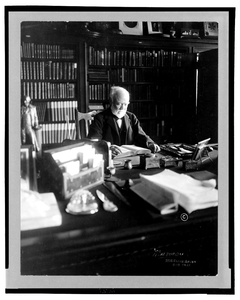 Andrew Carnegie, 1835-1919, half length, seated behind desk, looking down at book, facing right; shelves filled with books in background LCCN2005693059