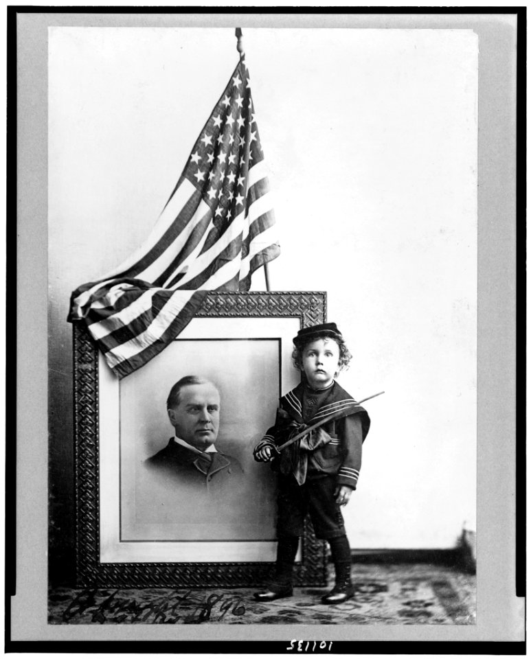 Boy in sailor uniform holding sword in front of flag-draped, head-and-shoulders, portrait of William McKinley LCCN90710001