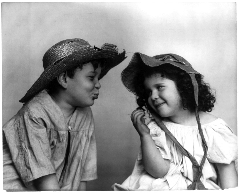 Boy wearing broken straw hat, with lips puckered, facing girl wearing bonnet LCCN96506058