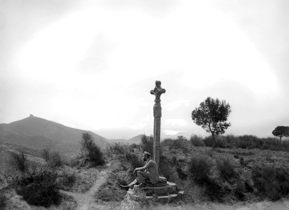 Creu de terme amb un senyor assegut i l'ermita de Sant Ramón al fons dalt del turóRestored