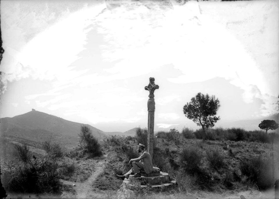 Creu de terme amb un senyor assegut i l'ermita de Sant Ramón al fons dalt del turó