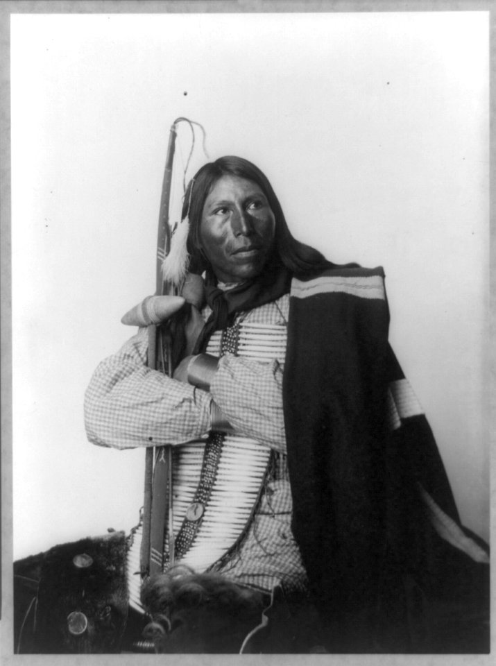 Distributor, Native American man, three-quarter length portrait, seated, facing right; arms folded, holding bow, arrow, and tomahawk; dressed in bone breast plate LCCN92508793